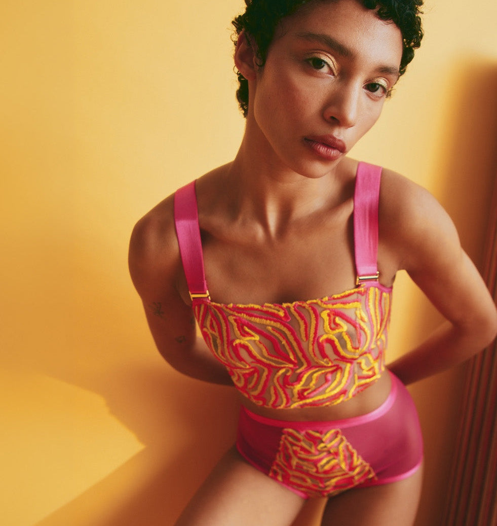 A model wearing a vibrant pink and yellow patterned two-piece swimsuit or bodysuit, posing against a warm yellow wall.